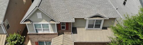 Two-story house with gray shingles, brick front, surrounded by greenery and a garage.