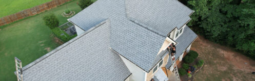 Roofing workers repair a house with a gray shingle roof amid a green lawn and trees.