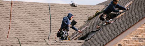 Two roofers work on a sloped roof, removing old shingles and preparing for repairs.