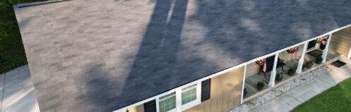 Aerial view of a single-story house with a dark shingled roof, green lawn, and porch decor.
