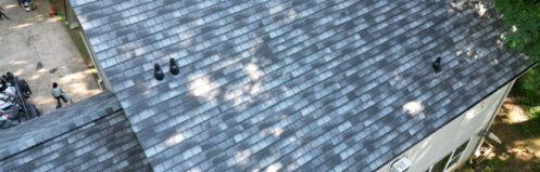 Aerial view of a gray shingled roof with vents, surrounded by trees and a driveway.