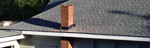 Roof with gray shingles and a brick chimney against green trees and blue sky.